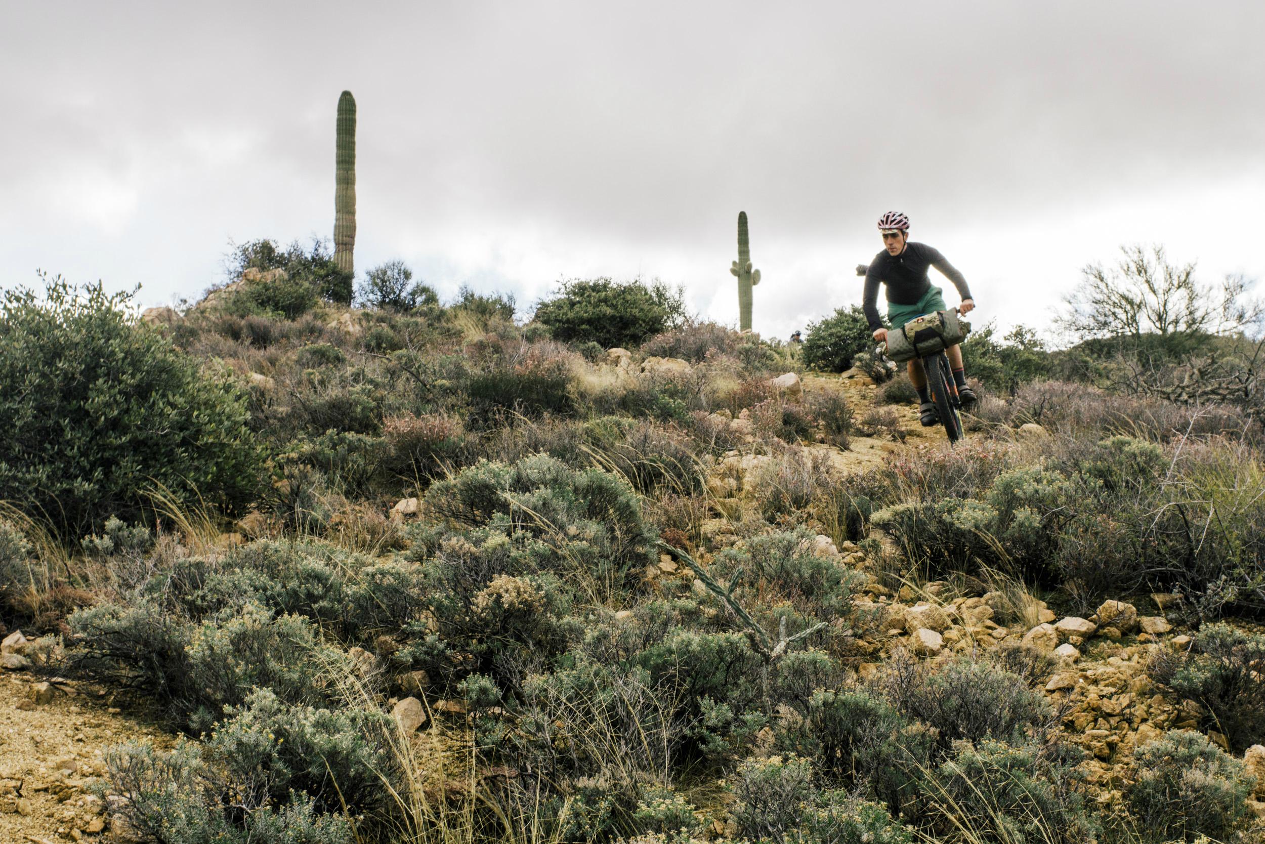 A cyclist navigating a rocky trail surrounded by desert vegetation and cacti under a cloudy sky. Arizona Trail mountain bike trail.