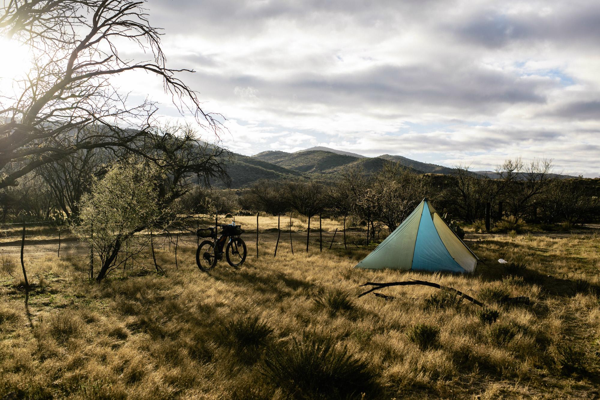 A blue and green tent set up in a grassy, open area surrounded by sparse trees and mountainous terrain in the background, with a bicycle parked nearby. The scene is illuminated by soft sunlight filtering through clouds, creating a tranquil outdoor camping atmosphere. Arizona Trail mountain bike trail.