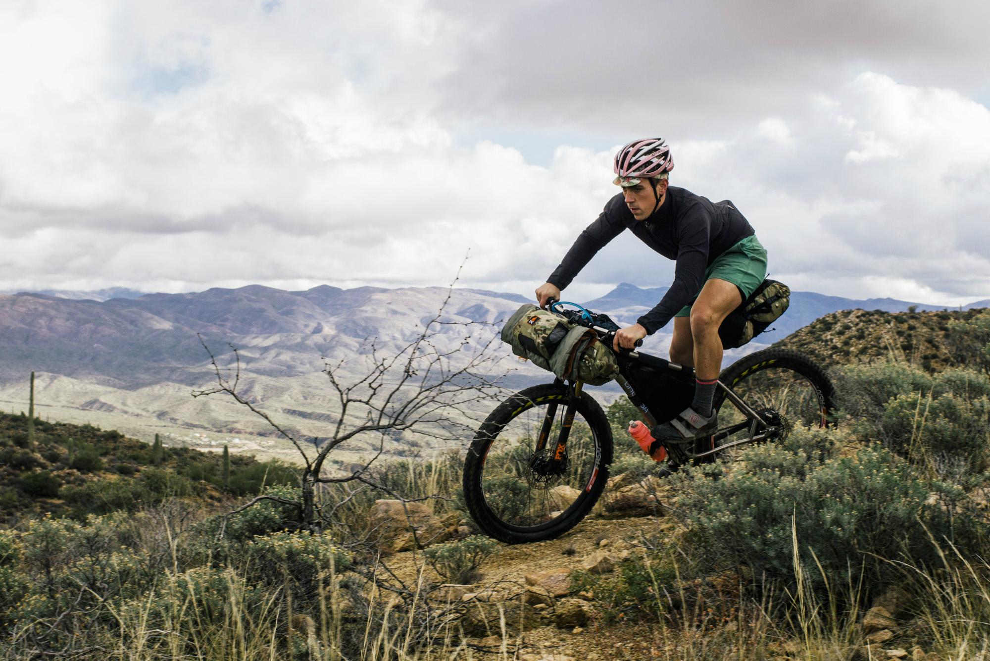 A cyclist riding a mountain bike on a rugged trail, equipped with bags for adventure, with a scenic view of mountains and cloudy skies in the background. Arizona Trail mountain bike trail.