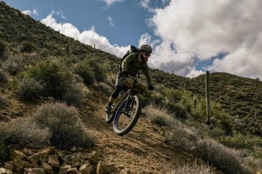 A mountain biker rides along a rocky trail in a desert landscape, surrounded by shrubs and cacti under a partly cloudy sky. The cyclist is wearing a helmet and sunglasses, with a backpack and gear attached to the bike. Arizona Trail mountain bike trail.