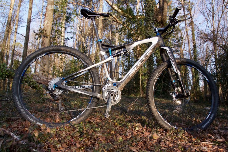 A mountain bike with a sleek design, featuring a silver and black frame, is positioned on a forest floor covered in leaves and small plants. The background includes tall trees and a clear sky, indicating a natural outdoor setting. The bike showcases thick tires and high-quality components, emphasizing its suitability for off-road riding.