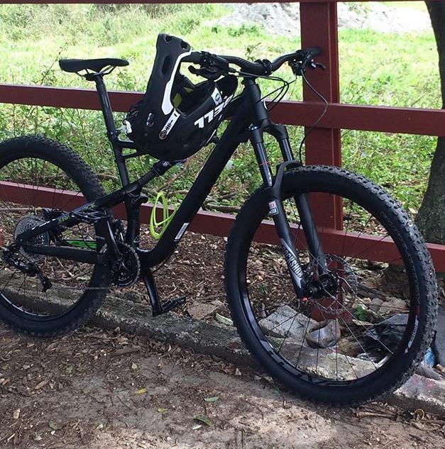 Specialized Camber 29: A black mountain bike with green accents, resting against a red wooden fence. A black helmet is secured on the bike's handlebars, and the background shows greenery and a natural setting.