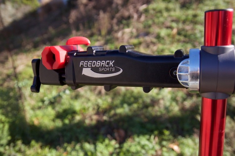 Close-up of a bike repair stand attachment featuring a black body with a red clamp. The attachment is marked with the text "FEEDBACK SPORTS" and surrounded by green grass in the background, indicating an outdoor setting.