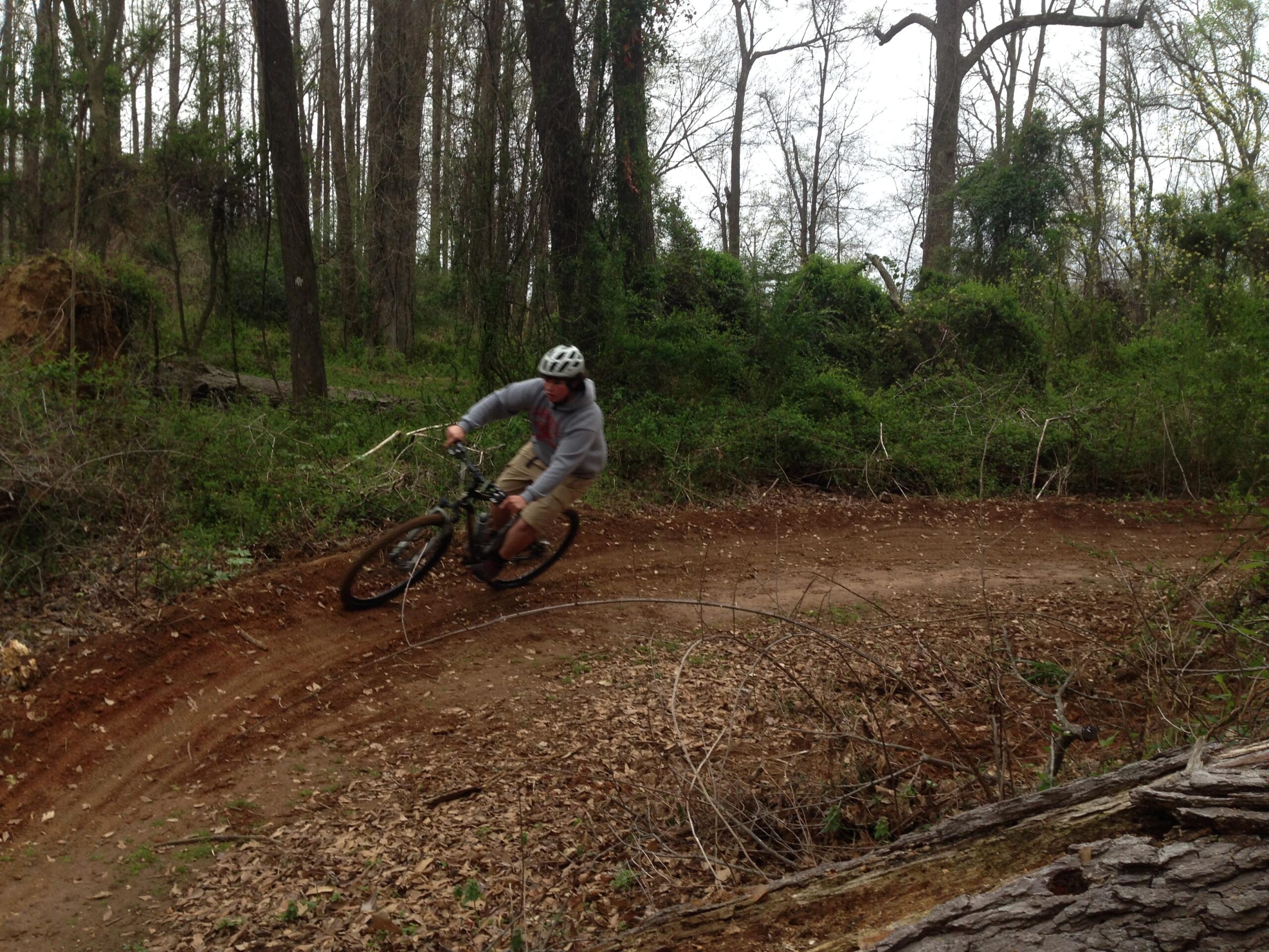 A person riding a mountain bike around a dirt curve on a wooded trail, surrounded by trees and greenery. The rider is wearing a helmet and a gray hoodie, navigating through an outdoor trail with scattered leaves on the ground. Spadra Creek Nature Trail mountain bike trail.