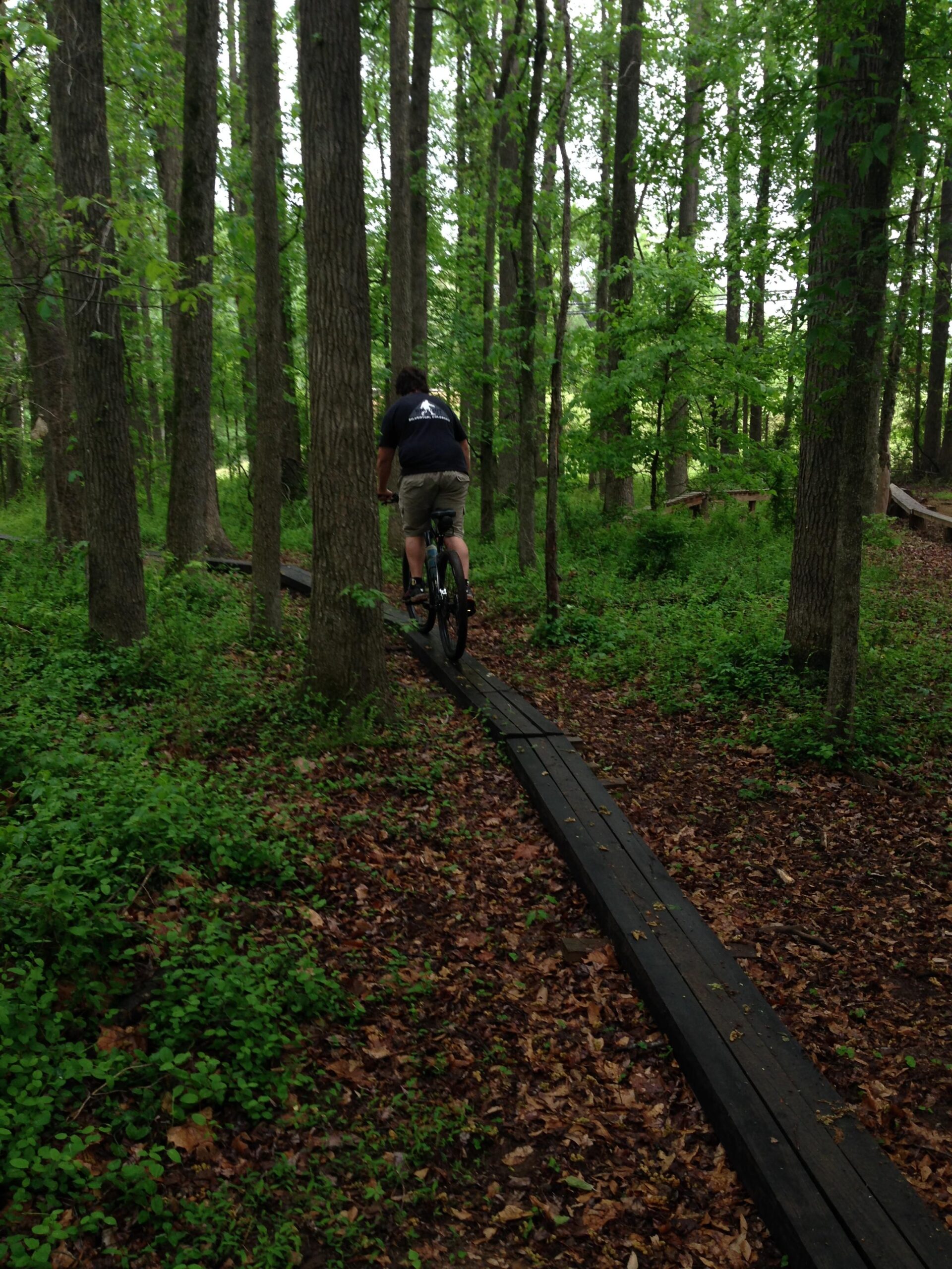 A person riding a mountain bike on a wooden plank trail through a lush green forest, surrounded by tall trees and foliage, with leaves scattered on the ground. Spadra Creek Nature Trail mountain bike trail.