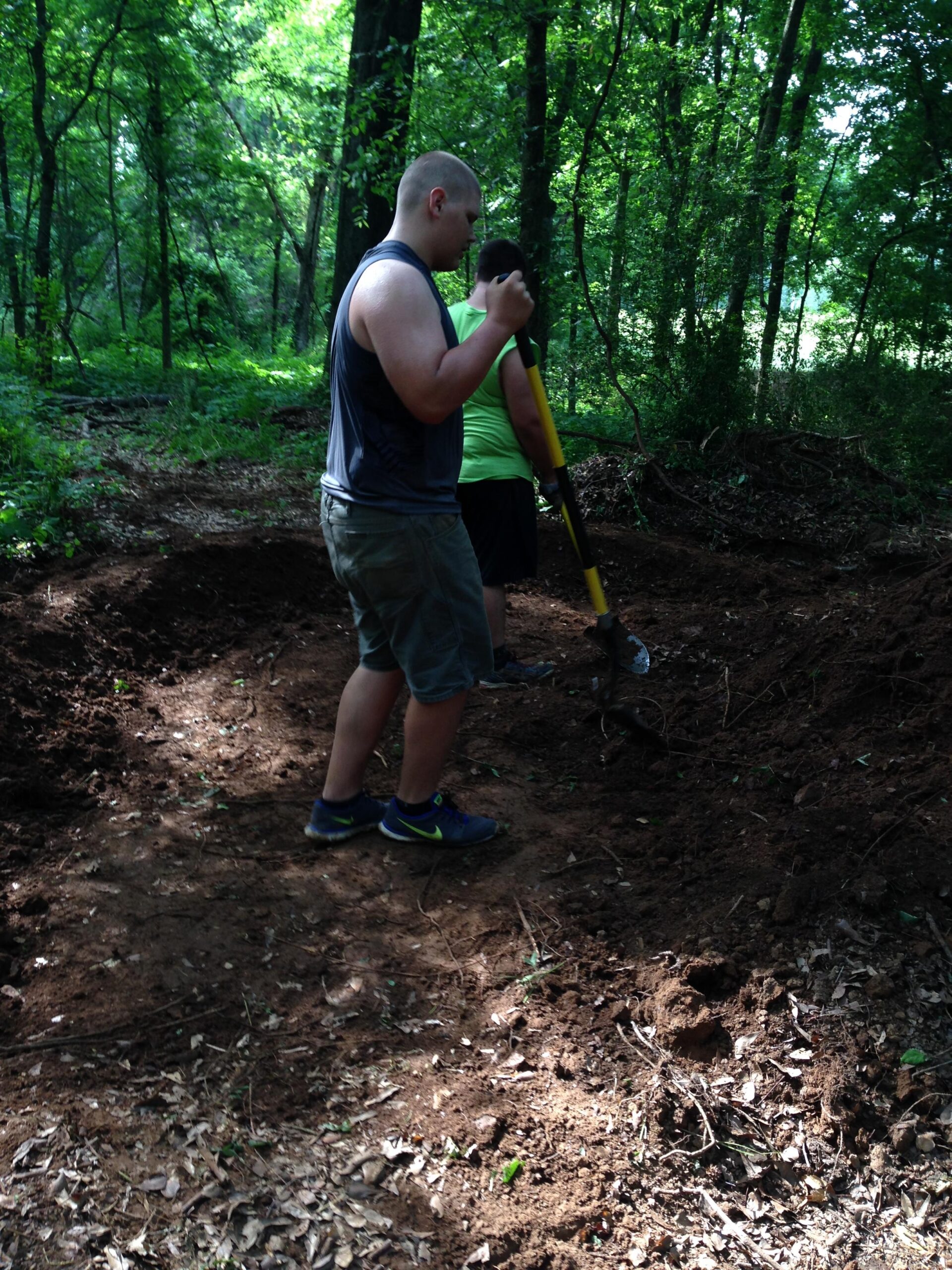 Two individuals are working together in a wooded area, digging in the ground. One person, wearing a sleeveless shirt and shorts, holds a shovel while looking down at the ground. The other person, dressed in a bright green shirt and shorts, stands nearby, actively participating in the digging effort. The surroundings are lush with greenery, including trees and leaves scattered on the ground. Spadra Creek Nature Trail mountain bike trail.