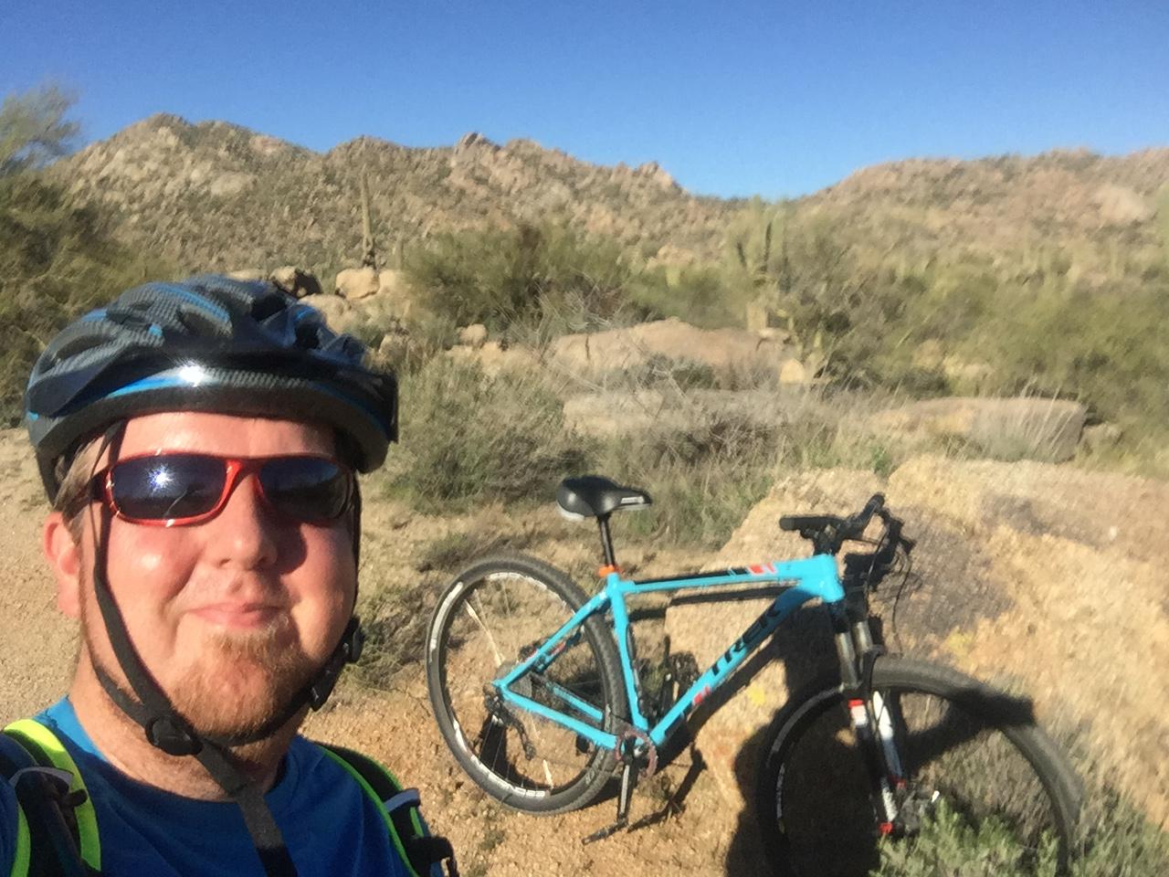 A person wearing a helmet and sunglasses smiles for a selfie while standing next to a blue mountain bike on a dirt trail in a desert landscape with rocky hills and sparse vegetation in the background. Brown's Ranch to Granite Mountain mountain bike trail.