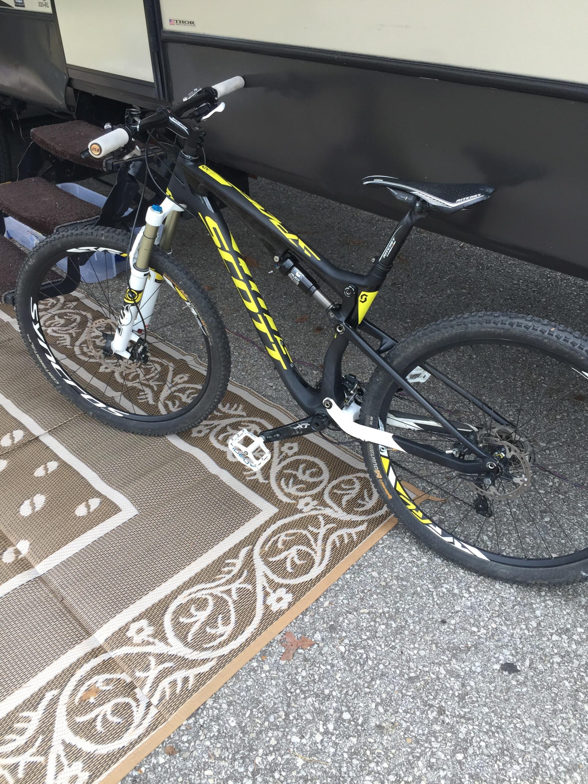 Scott Spark 700 RC: A black and yellow mountain bike is parked on a decorative mat outside an RV. The bike features adjustable handlebars and a sleek design, with visible suspension components. In the background, steps leading up to the RV are slightly visible, set against a gravel surface.