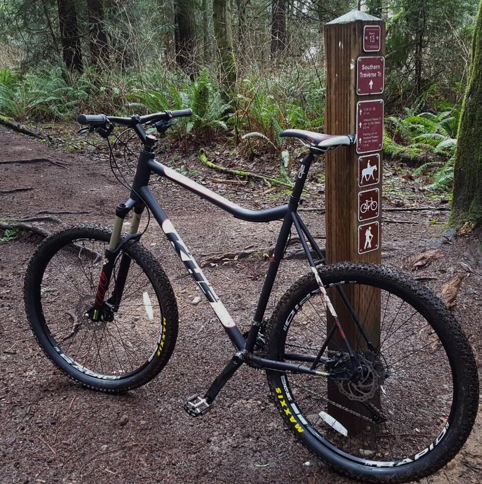 KHS BNT 29: A mountain bike leaning against a wooden trail sign in a forested area, surrounded by ferns and trees. The sign indicates trail directions and regulations for various activities, including biking and hiking. The ground is a mix of dirt and gravel, and the atmosphere appears lush and green.