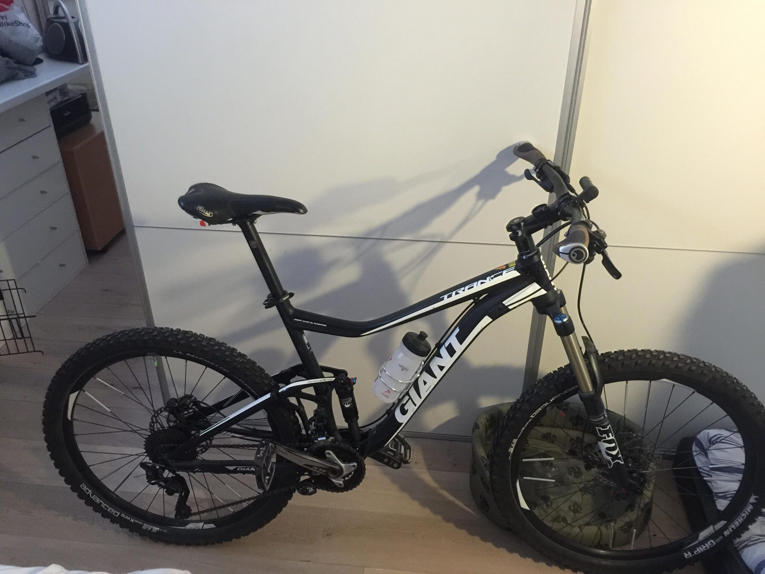 Giant Trance 27.5 2: A black and white mountain bike leaning against a light-colored wall. The bike features thick tires, a water bottle attached, and has a sporty design. In the background, there are storage drawers and items on shelves, indicating an indoor setting.