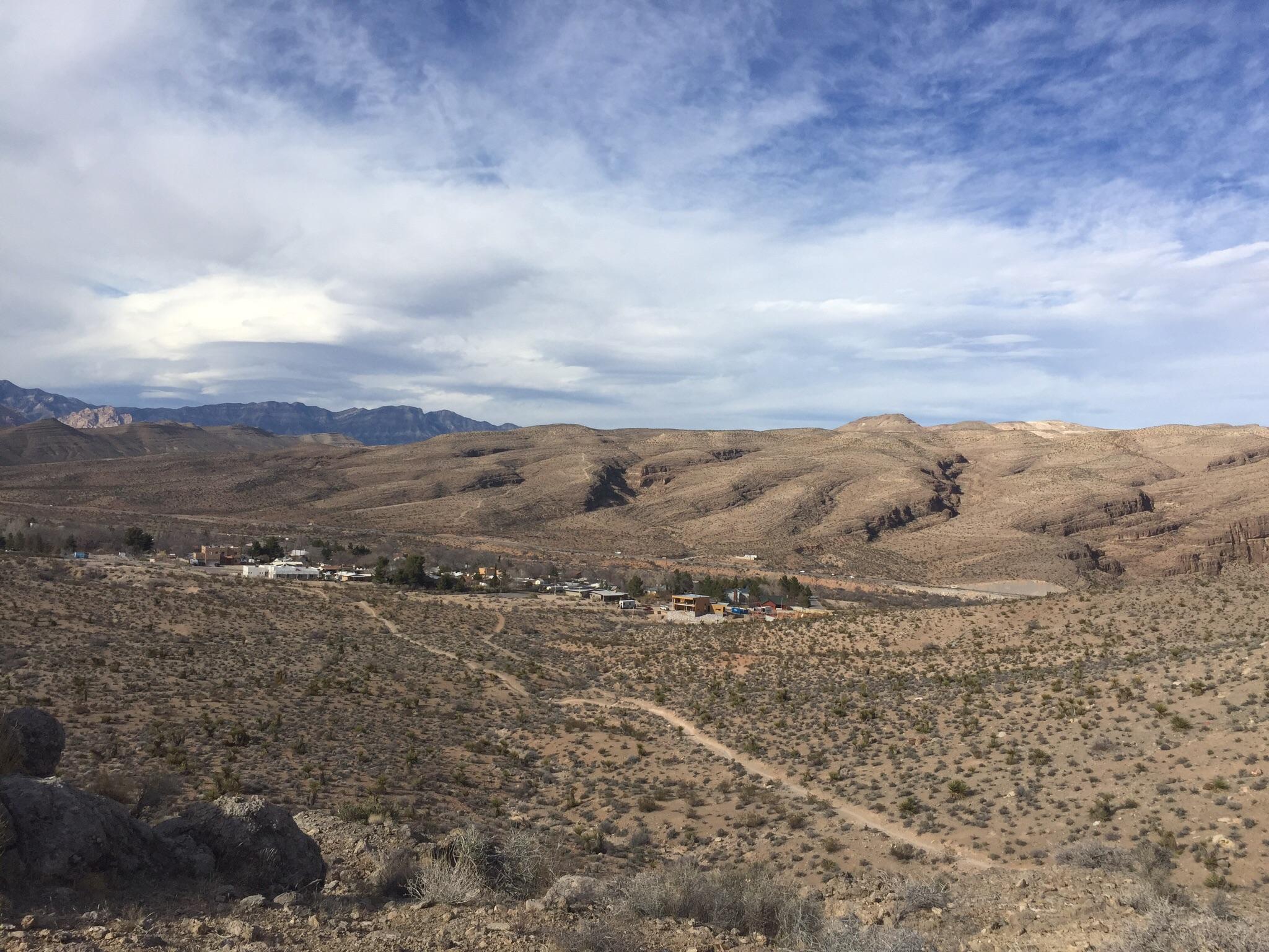 A panoramic view of a desert landscape featuring rolling hills and sparse vegetation, with a small settlement visible in the foreground. The background includes a range of mountains under a partly cloudy sky. The scene captures the rugged beauty of the arid environment. Blue Diamond mountain bike trail.