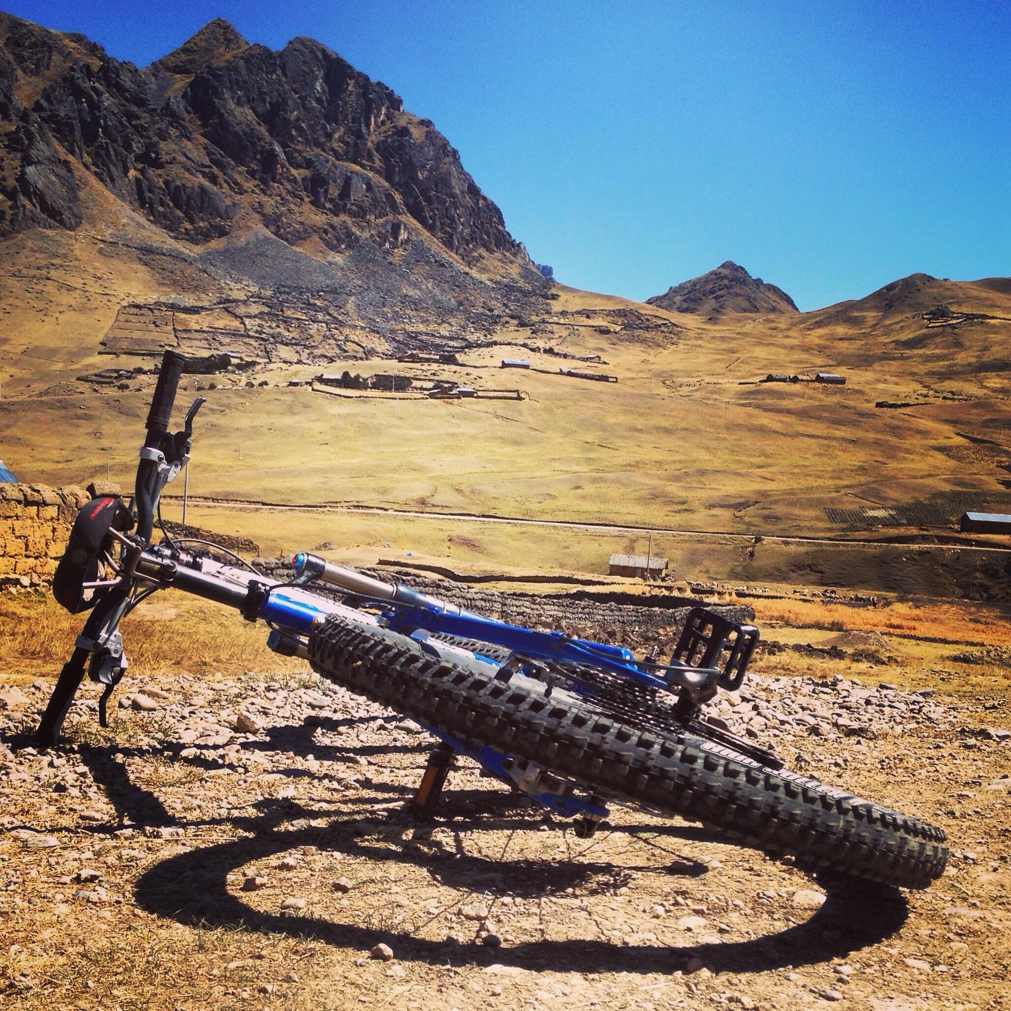 Rocky Mountain ETSX-70: A blue mountain bike rests on its side against a rocky, hilly terrain under a clear blue sky. In the background, there are distant mountains and patches of green fields, along with small structures scattered across the landscape. The image captures the serene beauty of an outdoor cycling environment.