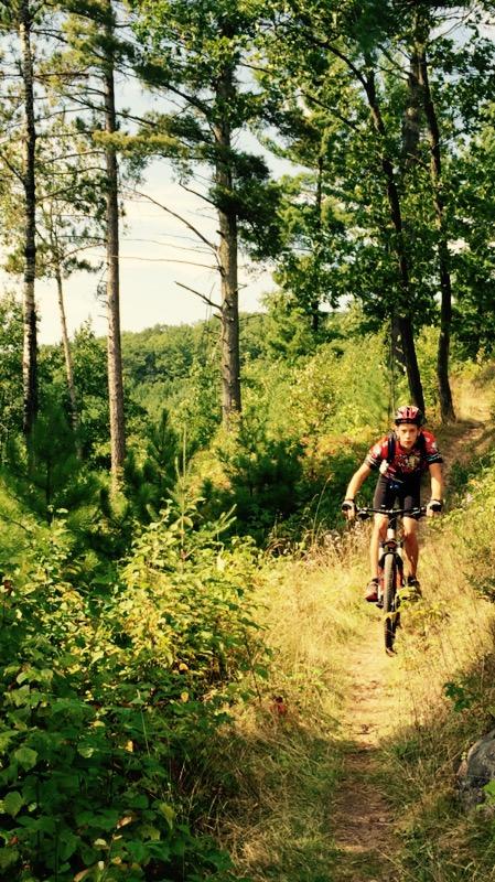 A mountain biker riding along a narrow, wooded trail surrounded by green trees and vegetation on a sunny day. Mud Lake mountain bike trail.