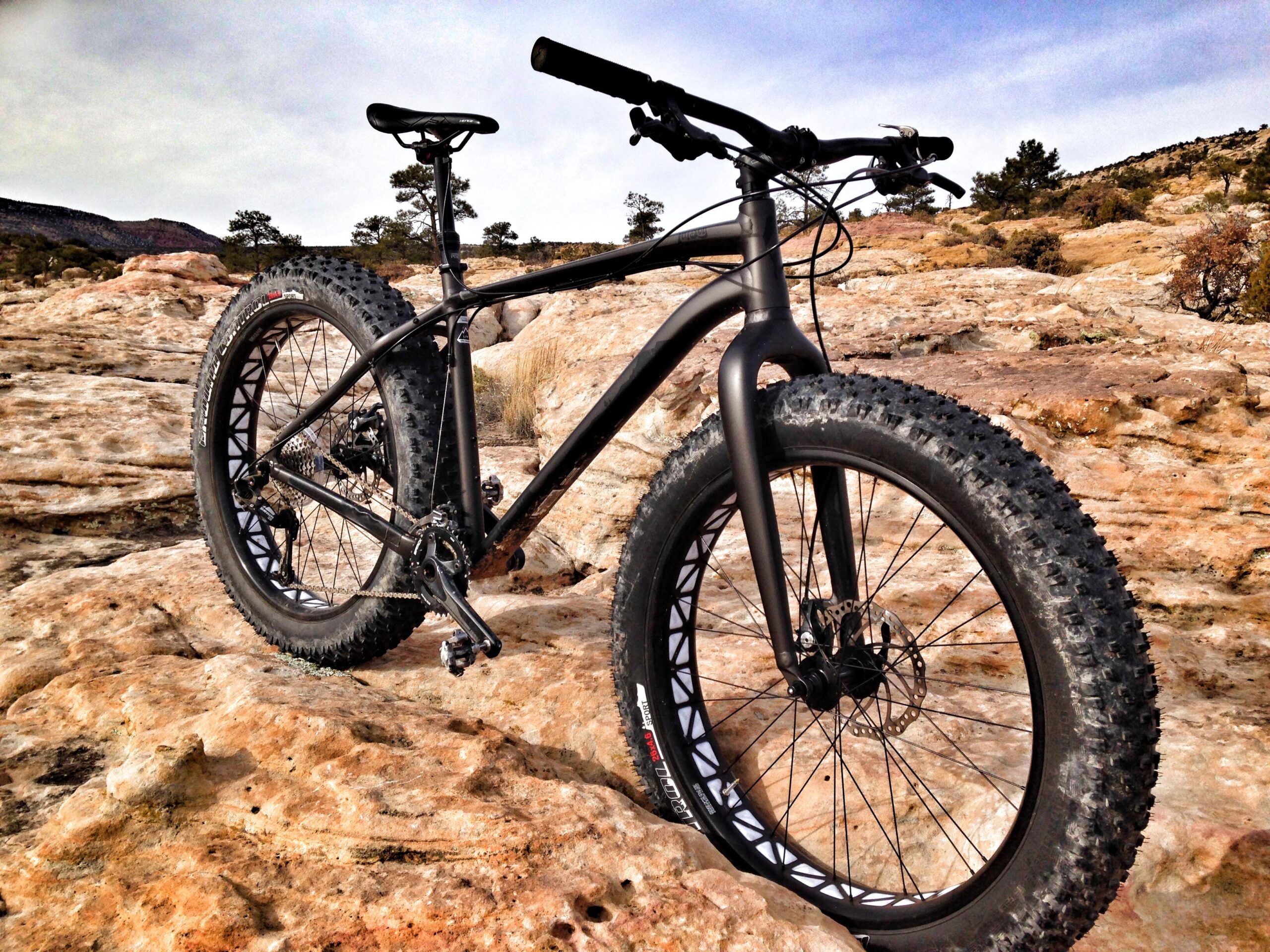 Specialized Fatboy: A black fat bike with wide, knobby tires rests on rocky terrain under a cloudy sky. The bike features a sleek frame and is set against a backdrop of natural landscape, showcasing its off-road capabilities.