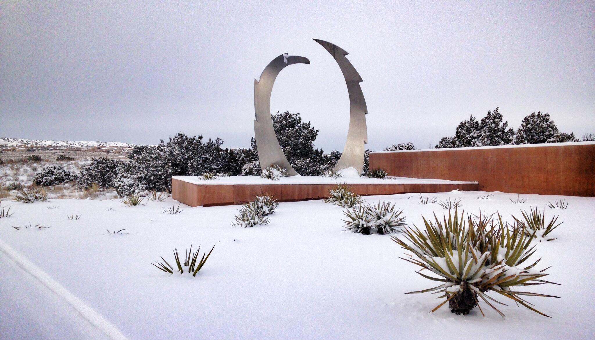 A snowy landscape featuring a modern metal sculpture shaped like two curved blades in the foreground. Surrounding the sculpture is a minimalist garden with patches of snow covering the ground and desert plants. In the background, a softly rolling hillside can be seen under overcast skies. Parkway Fatbike trail mountain bike trail.