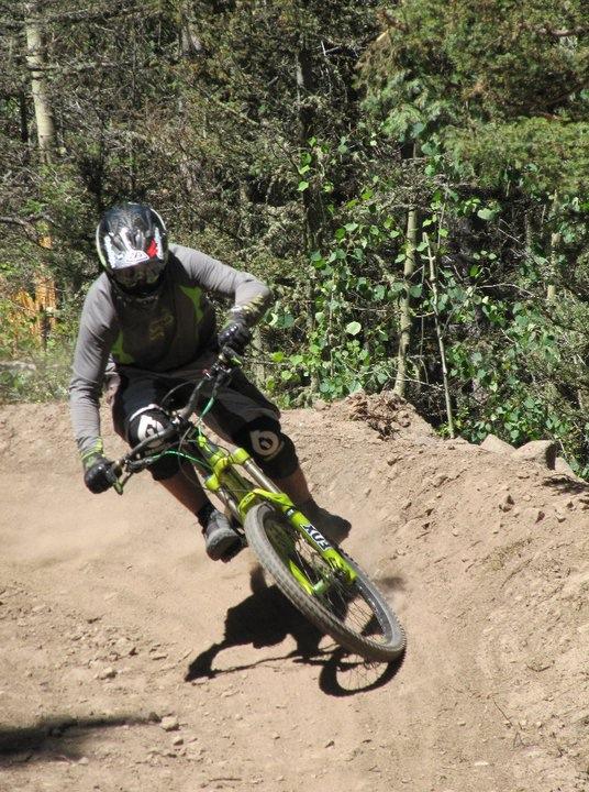 A mountain biker in protective gear leans into a turn on a dirt trail surrounded by trees, creating a cloud of dust. The bike is bright green with black accents, and the rider is focused on navigating the curve. Angel Fire Bike Park mountain bike trail.