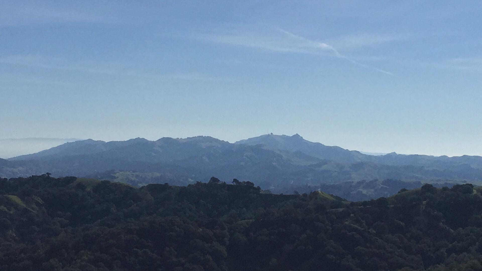 Expansive view of rolling green hills and distant blue mountains under a clear sky. The foreground features dense foliage, while the background displays a layered mountain range, creating a serene and natural landscape. Briones Regional Park mountain bike trail.