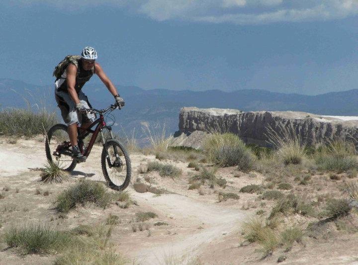 Specialized Enduro SL Pro: A mountain biker navigating a dirt trail on a sunny day, with a rocky cliff and distant mountains in the background. The biker is wearing a helmet and a backpack, showcasing a blend of vegetation and dry terrain.