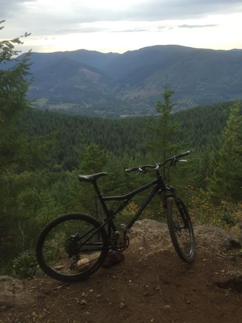 Santa Cruz Blur: A black mountain bike parked on a rocky outcrop, overlooking a lush green valley filled with trees and distant mountains under a cloudy sky.