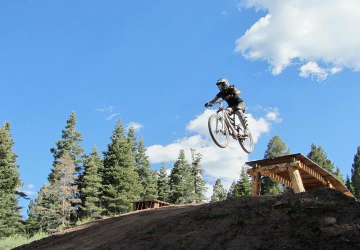 A mountain biker in a helmet is performing a jump off a dirt ramp, airborne above a wooded area with blue skies and clouds in the background. Angel Fire Bike Park mountain bike trail.
