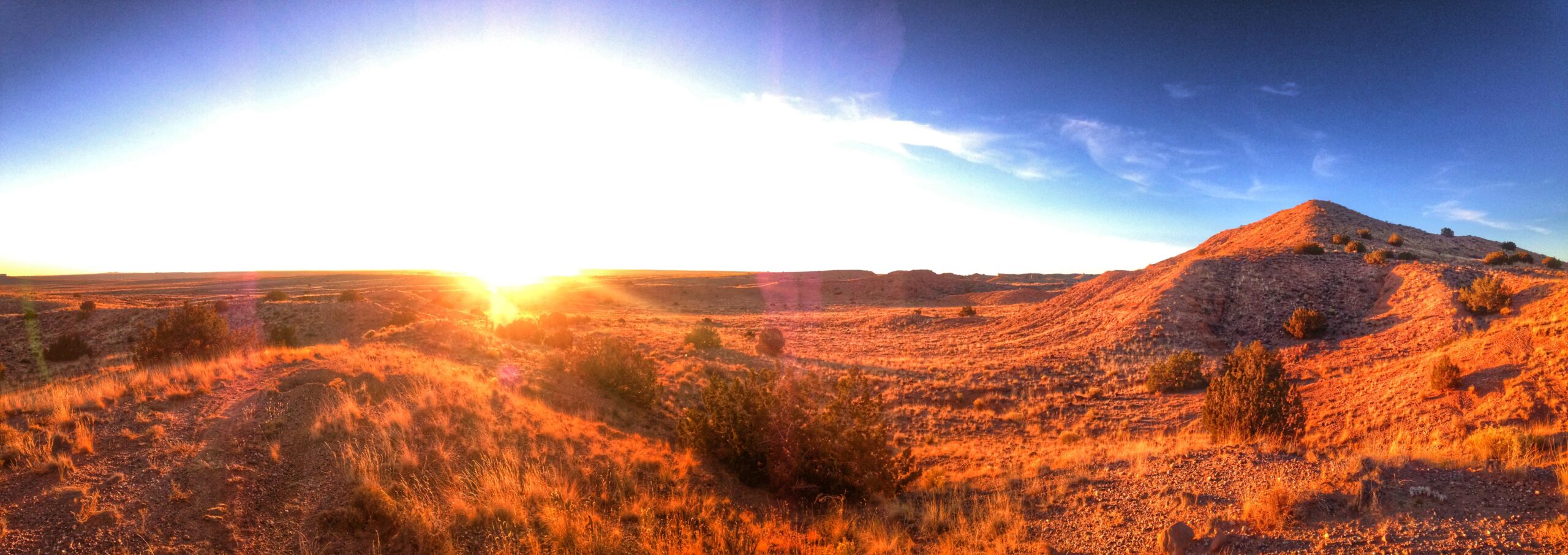 A panoramic view of a sunlit desert landscape during sunrise, showcasing rolling hills and sparse vegetation. The sky transitions from deep blue at the top to bright orange and yellow near the horizon, with rays of sunlight illuminating the reddish-brown terrain and scattered bushes in the foreground. Parkway Fatbike trail mountain bike trail.