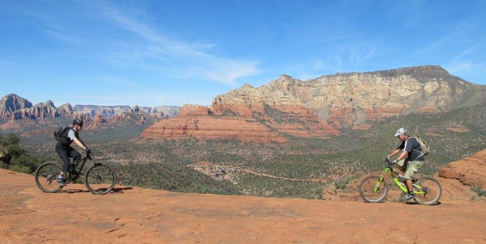 Two mountain bikers ride along a rocky trail with a backdrop of red rock formations and a clear blue sky. One biker is wearing a black helmet and backpack, while the other sports a green bike and a white helmet. The landscape features rugged cliffs and vegetation typical of a desert environment. Bell Rock Area Trails mountain bike trail.