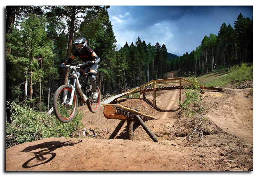 A mountain biker in protective gear jumps over a wooden ramp on a dirt trail, surrounded by dense trees and a cloudy sky. Angel Fire Bike Park mountain bike trail.