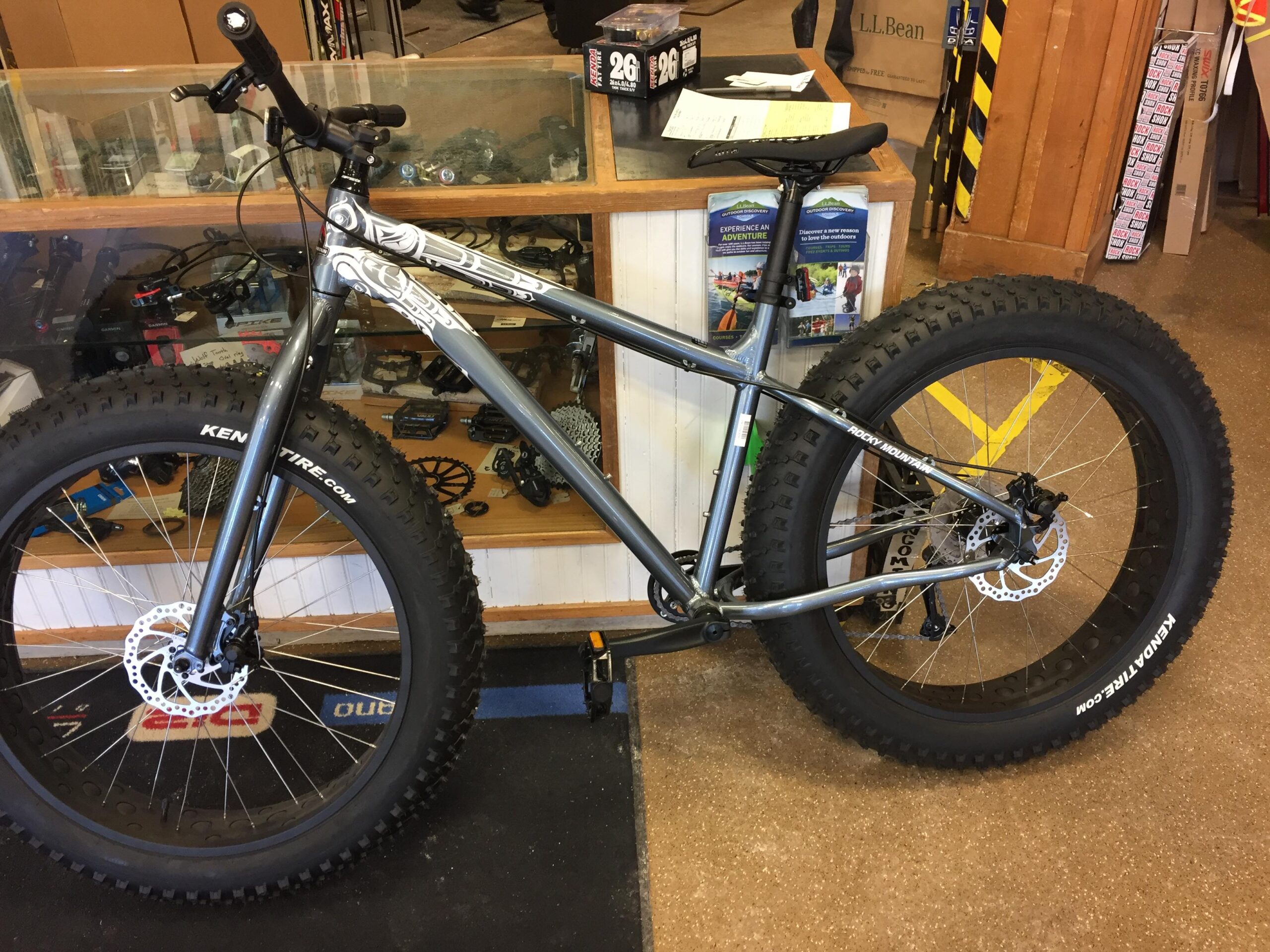 Rocky Mountain Blizzard: A gray fat bike with large, knobby tires displayed in a shop. The bike features a sleek design with prominent branding and a disc brake on the front wheel. The background includes a display case with various bike components visible.