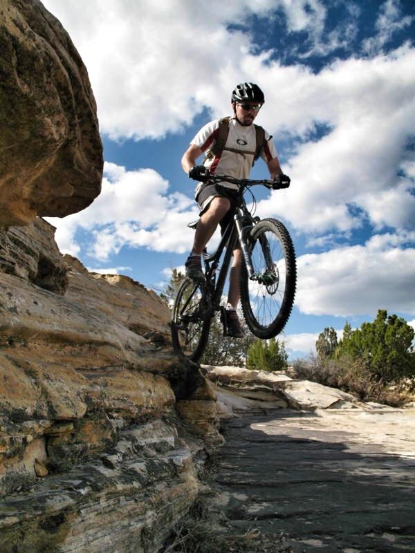 Specialized Stumpjumper Comp: A mountain biker performs a jump off a rocky ledge, with a blue sky and fluffy clouds in the background. The biker is wearing a helmet and riding gear, showcasing an active outdoor lifestyle in a rugged terrain.