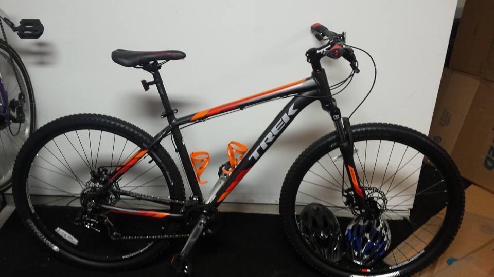 Trek Marlin 5 29er: A black and orange Trek mountain bike is leaning against a white wall. The bike features thick tires and a comfortable seat, with visible gear components and a handlebar with grips. In the background, there are cardboard boxes and another bike partially visible.