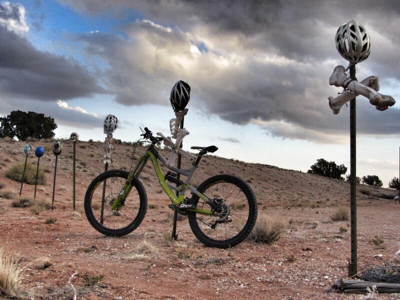 A mountain bike stands on a dusty landscape decorated with various helmeted skeleton sculptures. Each skeleton is mounted on a pole, showcasing different helmet designs against a backdrop of dramatic clouds and sparse vegetation. The scene conveys a mix of whimsy and caution, highlighting the importance of safety in biking.