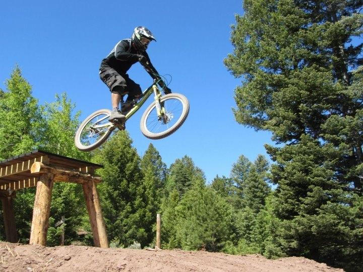 A mountain biker performing a jump off a wooden ramp, surrounded by trees under a clear blue sky. Angel Fire Bike Park mountain bike trail.