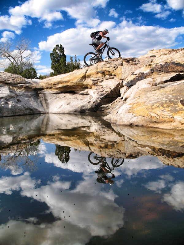 A mountain biker navigating a rocky ledge above a calm body of water, reflecting the blue sky and fluffy white clouds. Green trees are visible in the background, emphasizing the natural landscape.