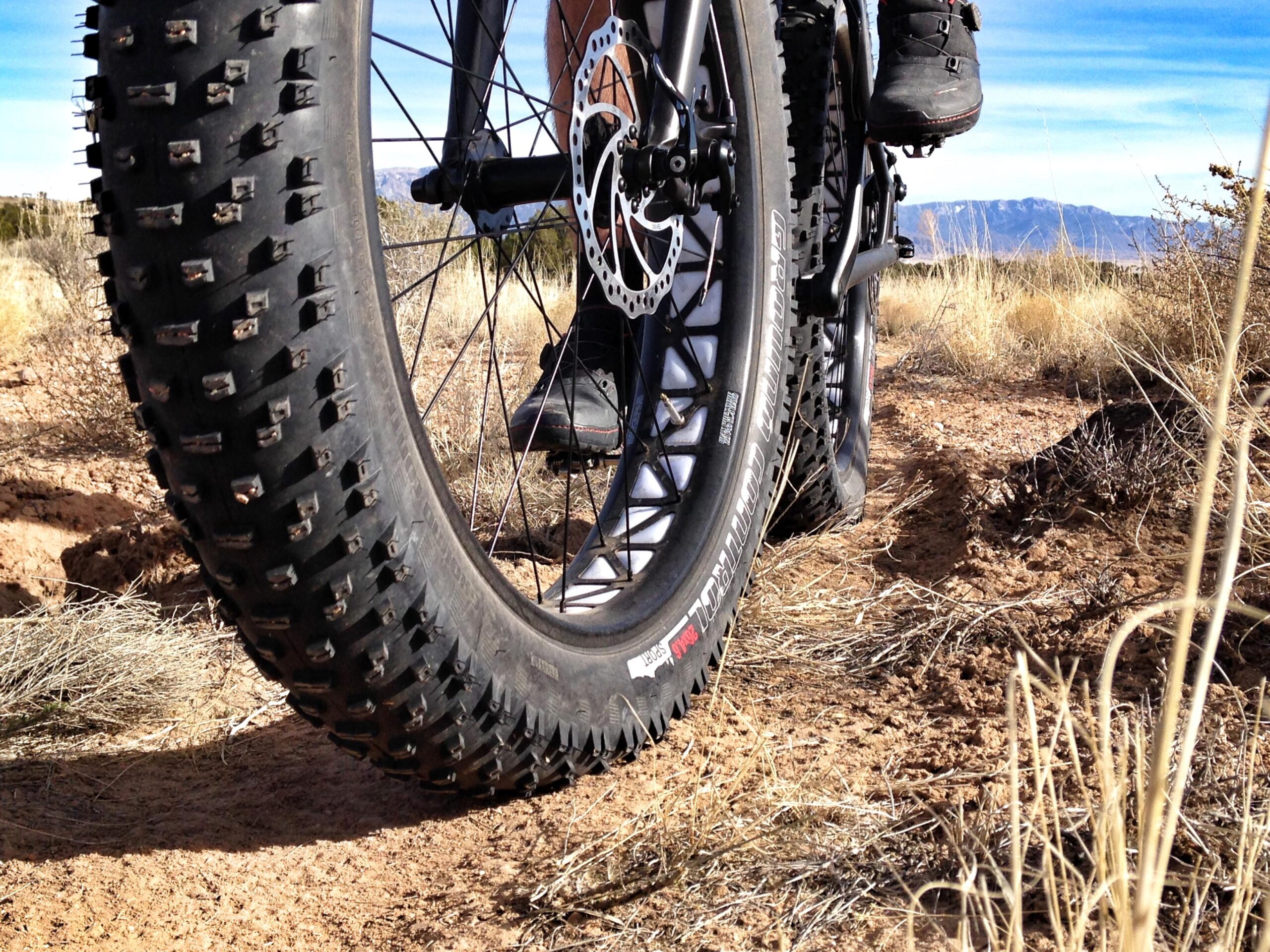 A close-up view of a fat bike tire on a sandy trail, with a rider's foot resting on the pedal. The tire features deep, knobby treads designed for off-road traction, surrounded by dry grasses and a clear blue sky in the background. Parkway Fatbike trail mountain bike trail.