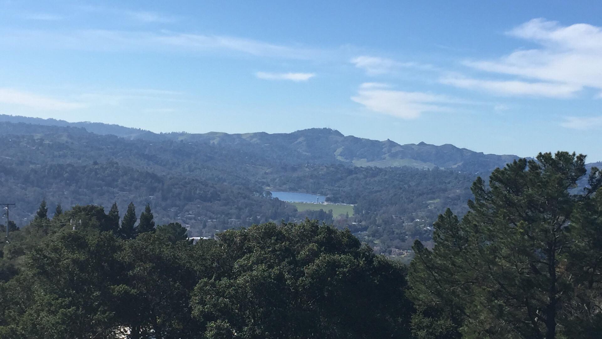 A panoramic view of a mountainous landscape featuring rolling hills and a blue lake nestled among the greenery, under a clear blue sky with a few clouds. Briones Regional Park mountain bike trail.