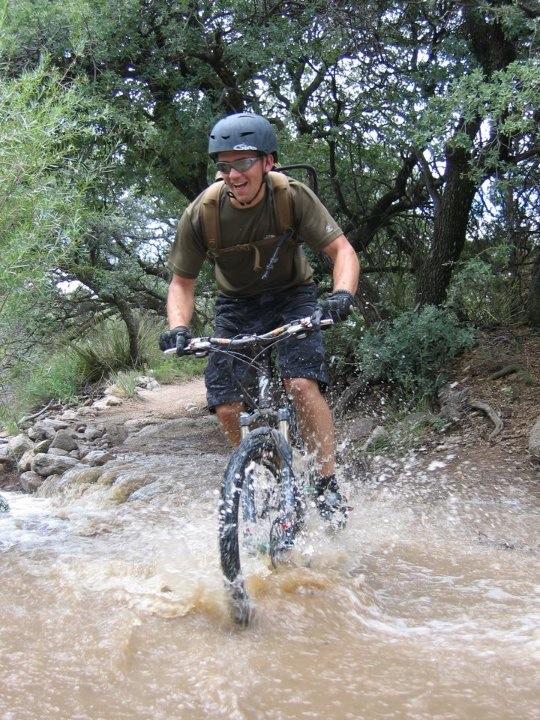 A cyclist wearing a helmet and sunglasses rides through a muddy puddle on a rocky trail surrounded by greenery, splashing water as he navigates the terrain.