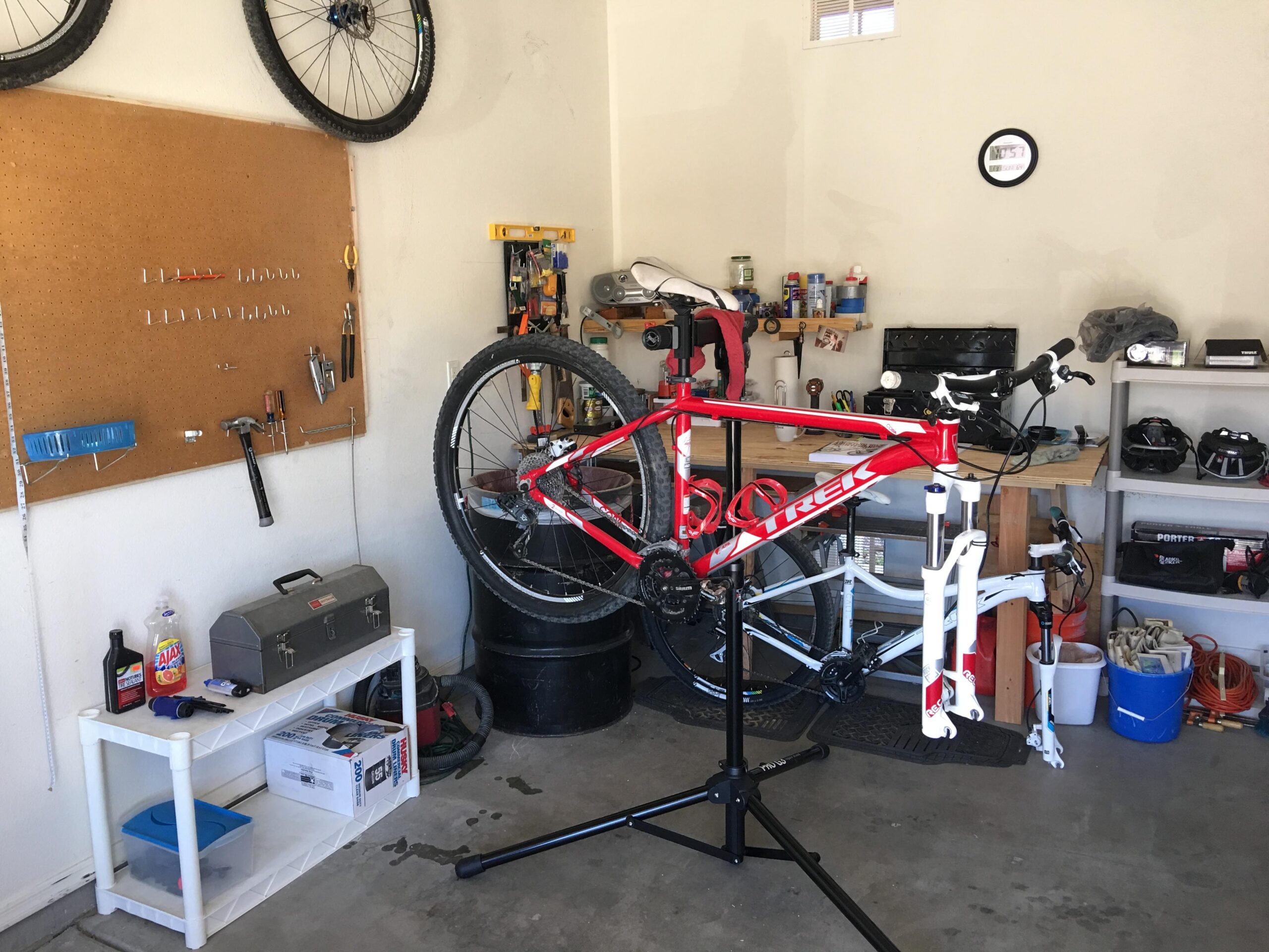 Trek Cobia: Alt text: A cluttered garage workspace featuring a red Trek bicycle mounted on a repair stand. The wall displays a pegboard with various tools hanging, and there are shelves with helmets, bottles, and a toolbox. A white table holds cleaning supplies and bike accessories, while a black container and additional items are visible in the background.