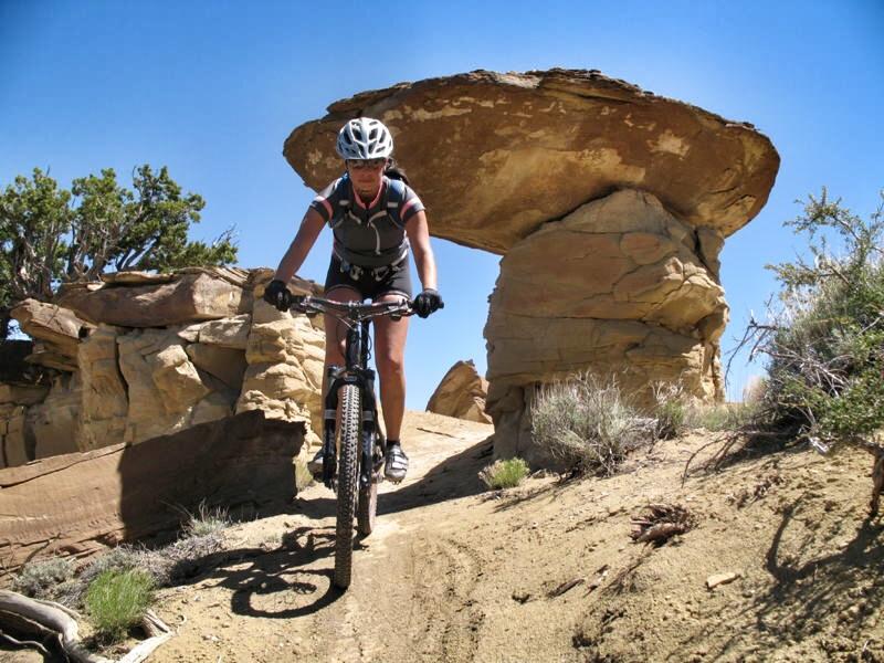 Specialized S-Works Stumpjumper FSR Carbon: A mountain biker navigating a dirt trail surrounded by unique rock formations under a clear blue sky. The biker, wearing a helmet and cycling gear, is focused on maneuvering around the terrain.