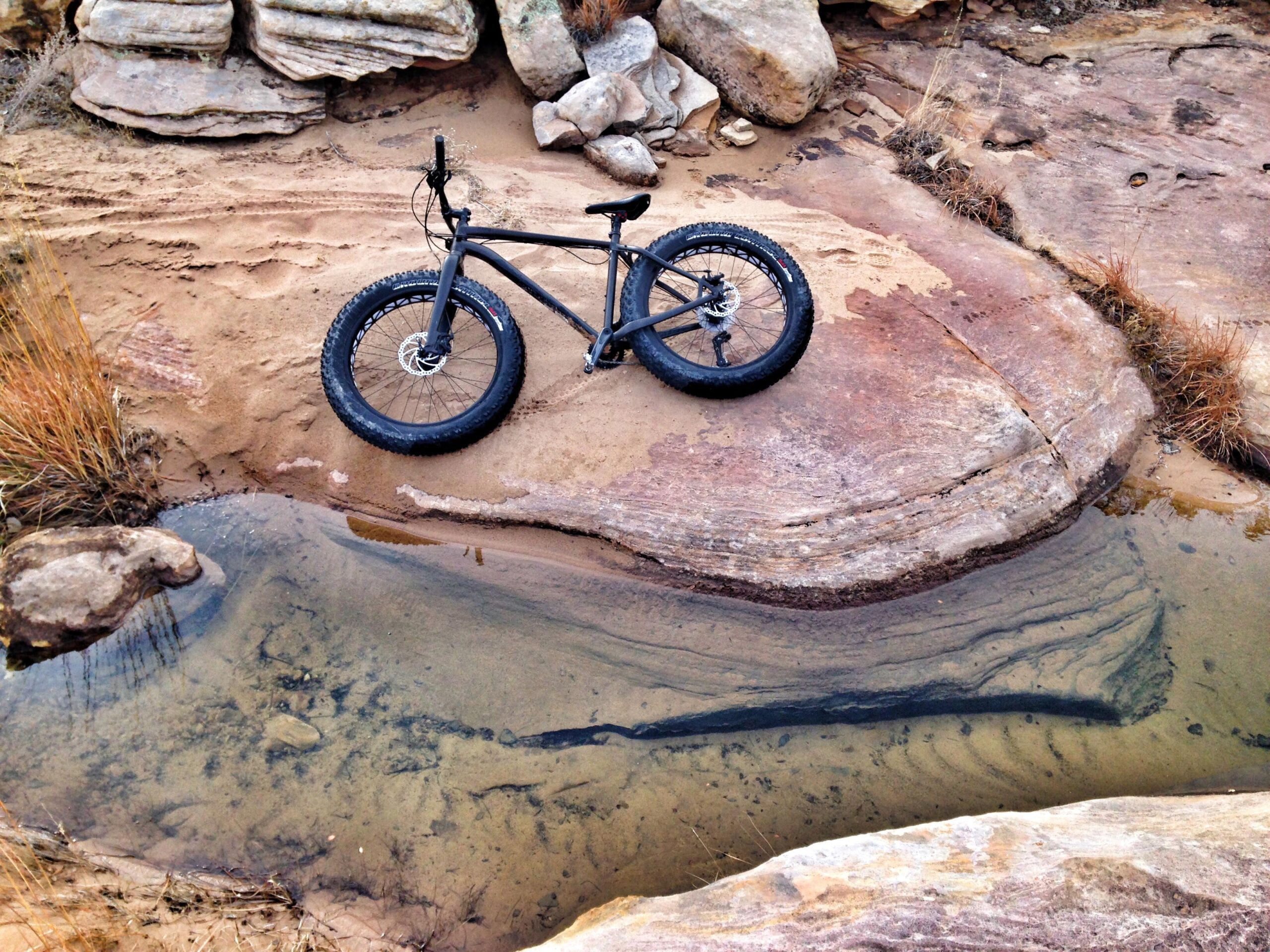 Specialized Fatboy: A black fat tire bike resting on a rocky terrain near a shallow pool of clear water. Surrounding the bike are various large stones and patches of grass, with sandy ground visible in the background.