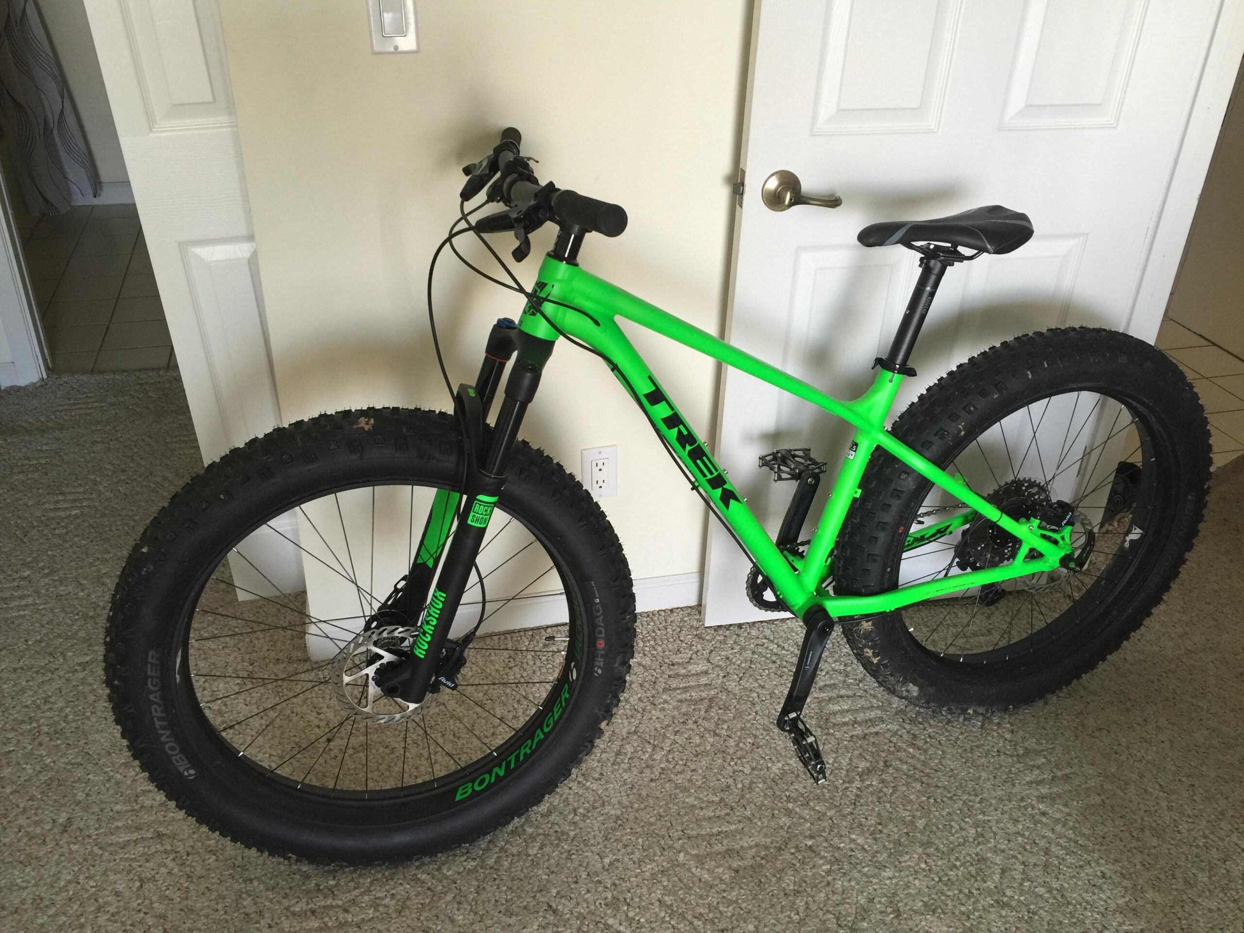 Trek EX 8: A bright green fat tire bike leaning against a wall with a closed door in the background. The bike features wide, knobby tires suitable for off-road riding and has a sleek design with a black seat and handlebars. The floor is covered in carpet, and there are tiled flooring visible in the background.