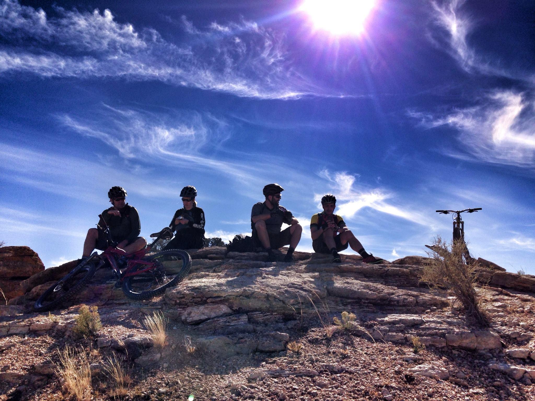 Four mountain bikers resting on a rock formation under a bright sun with a clear blue sky and wispy clouds. Their bikes are nearby, and they appear to be enjoying a break in a scenic outdoor setting. White Ridge Bike Trails mountain bike trail.