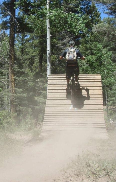 A mountain biker riding off a wooden ramp in a forested area, kicking up dust as they ascend into the air. Tall trees surround the trail, creating a natural backdrop. Angel Fire Bike Park mountain bike trail.