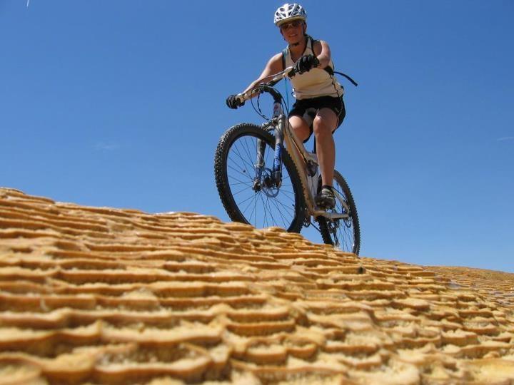 A person riding a mountain bike over a textured, sandy surface under a clear blue sky. The rider is wearing a helmet and athletic gear, focused on navigating the terrain. White Ridge Bike Trails mountain bike trail.