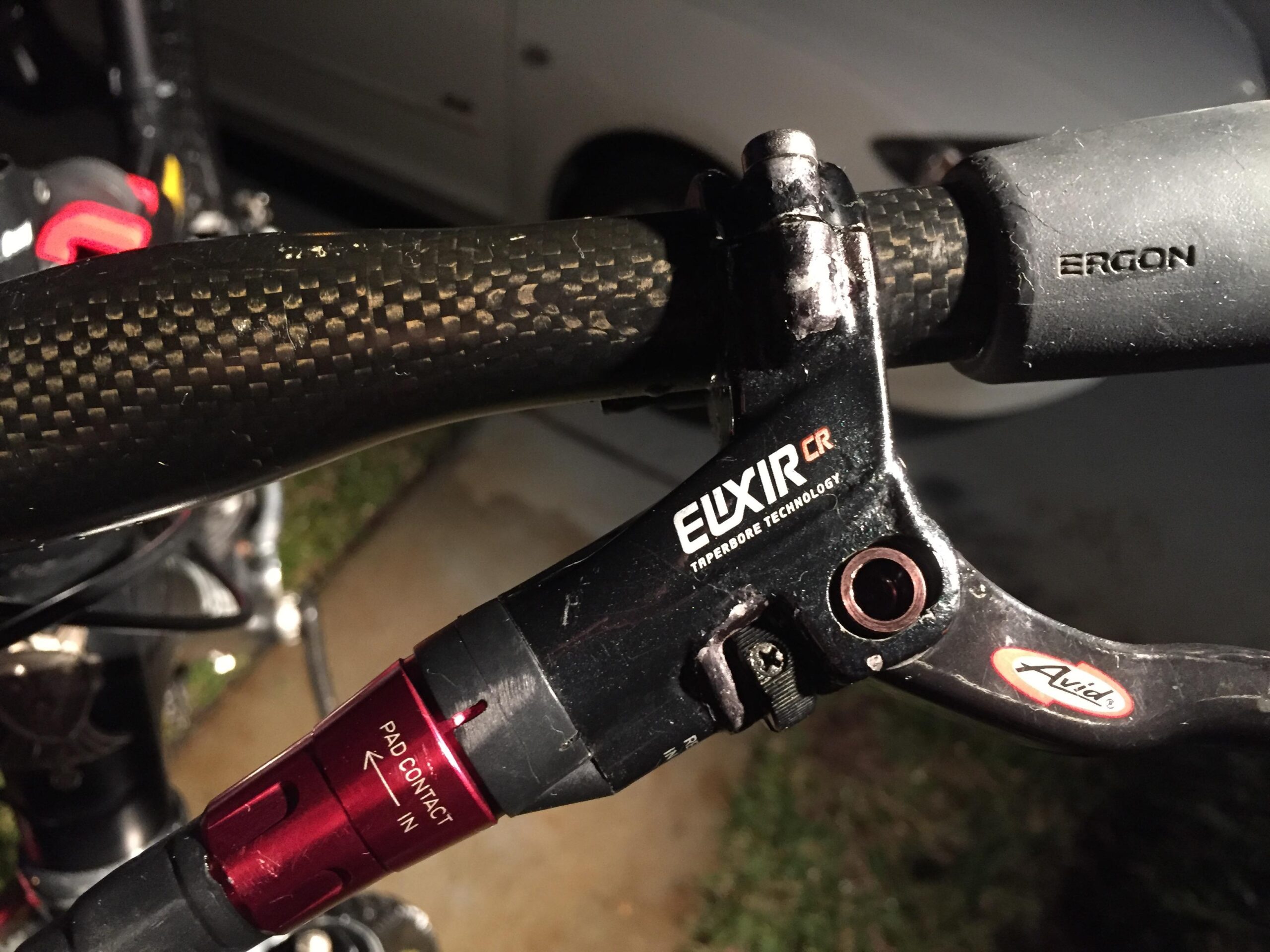 Voodoo Cycles Canzo 29er F/S: Close-up image of a bicycle brake lever mounted on a handlebar, featuring a carbon fiber grip and labeled with "ELIXIR CR" and "Avid." The image highlights a red adjustment component for pad contact, set against a dimly lit background.
