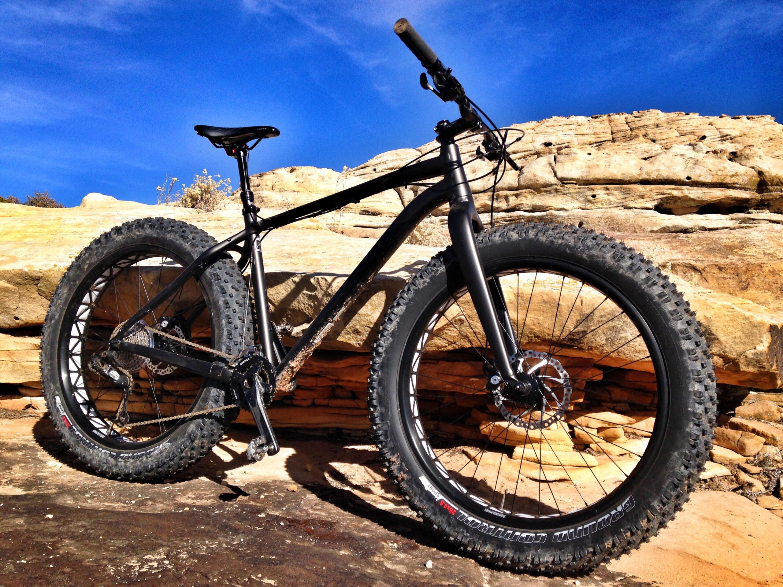 Specialized Fatboy: A black fat bike with large, knobby tires is leaning against a rocky outcrop under a clear blue sky. The bike's frame is sleek and modern, showcasing its sturdy design, while the background features rugged rock formations and earthy tones.