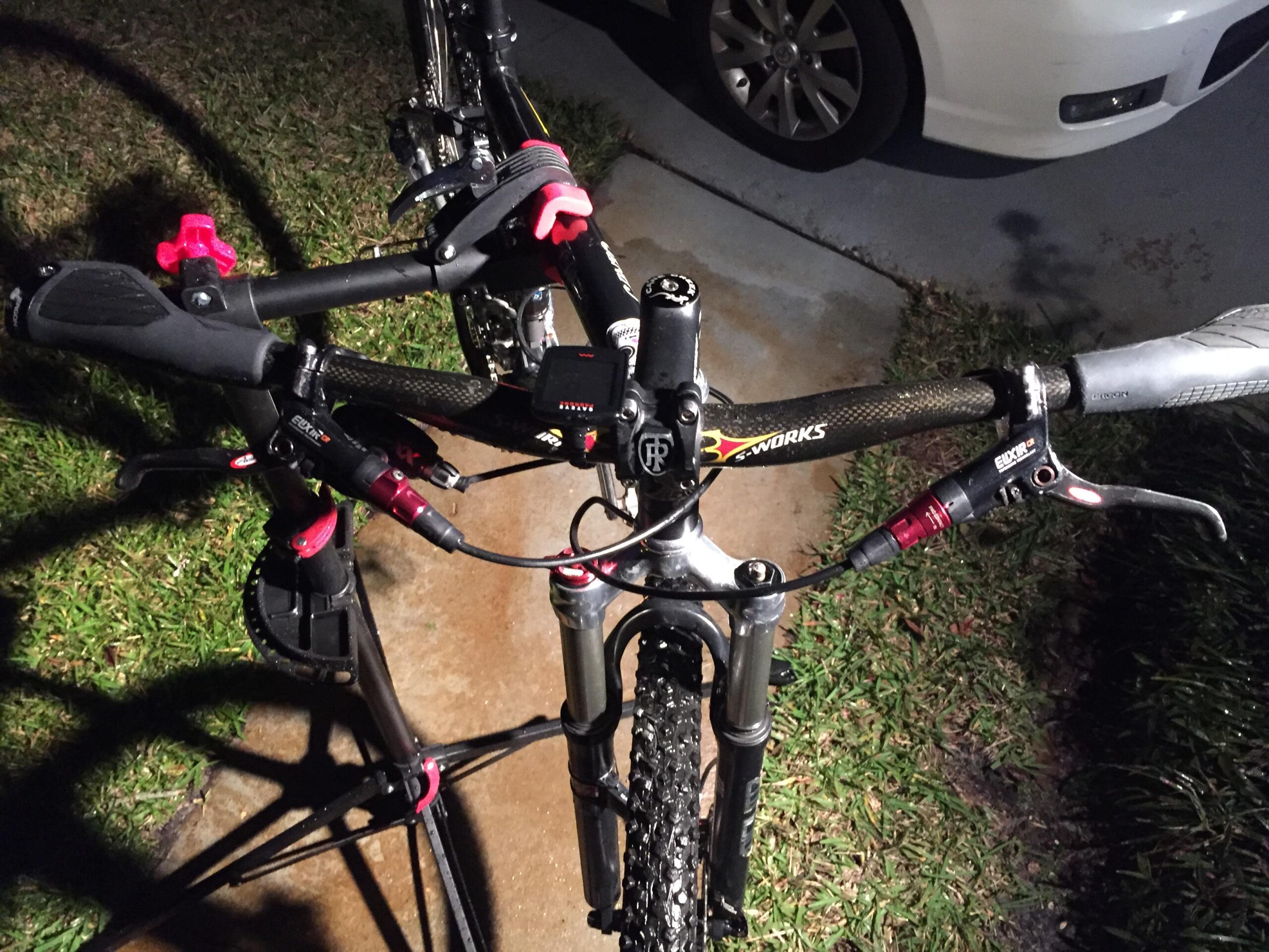 Voodoo Cycles Canzo 29er F/S: Close-up view of a mountain bike's handlebars at night, featuring prominent red and black components, including brake levers, shifters, and a digital display. The bike is positioned on a concrete surface with green grass visible in the background.