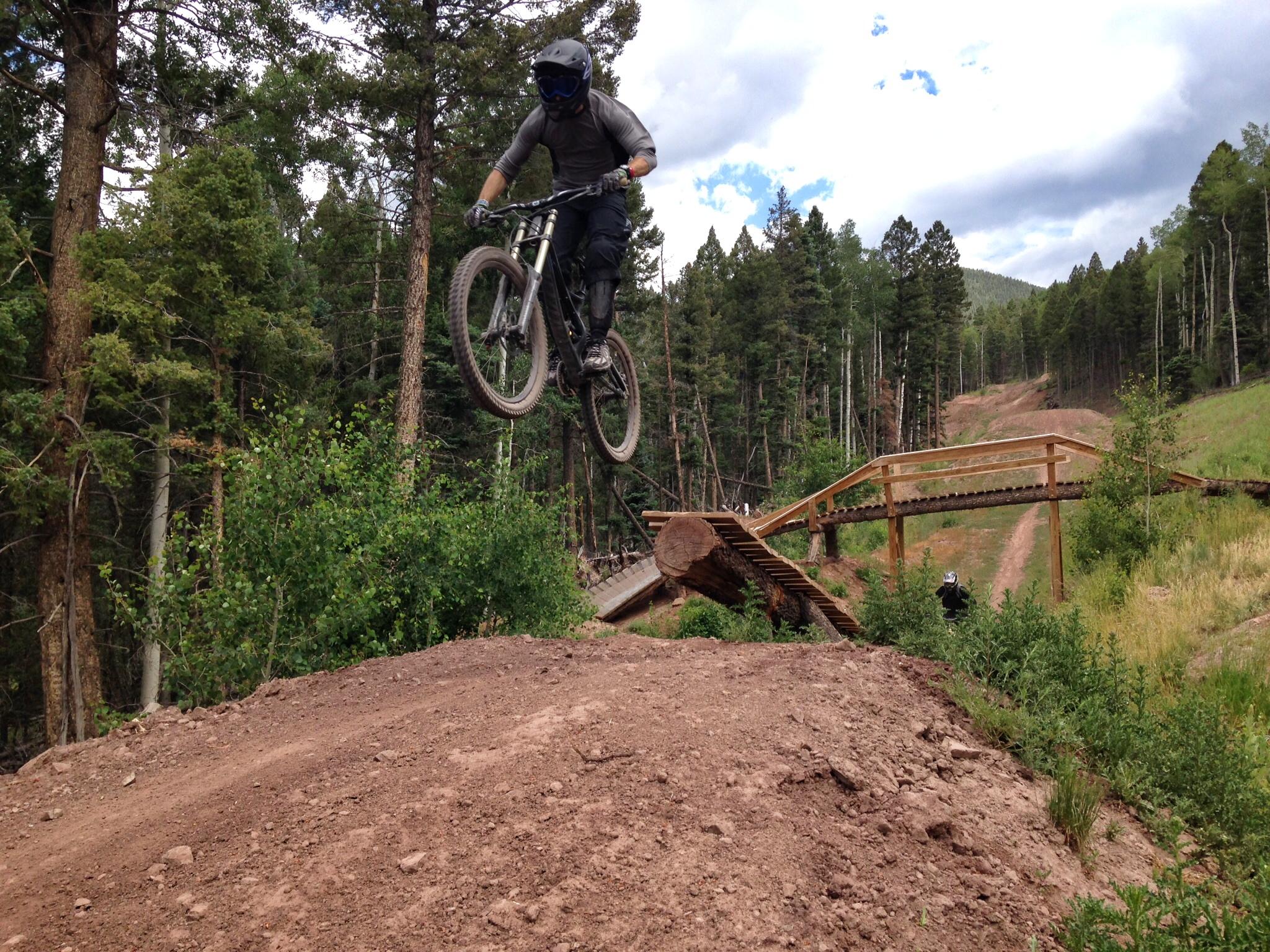 A mountain biker wearing a helmet and protective gear is seen jumping off a dirt ramp in a forested area, with trees in the background and a wooden bridge in the distance. The biker is mid-air, showcasing a dynamic riding pose against a backdrop of green foliage and a winding trail. Angel Fire Bike Park mountain bike trail.