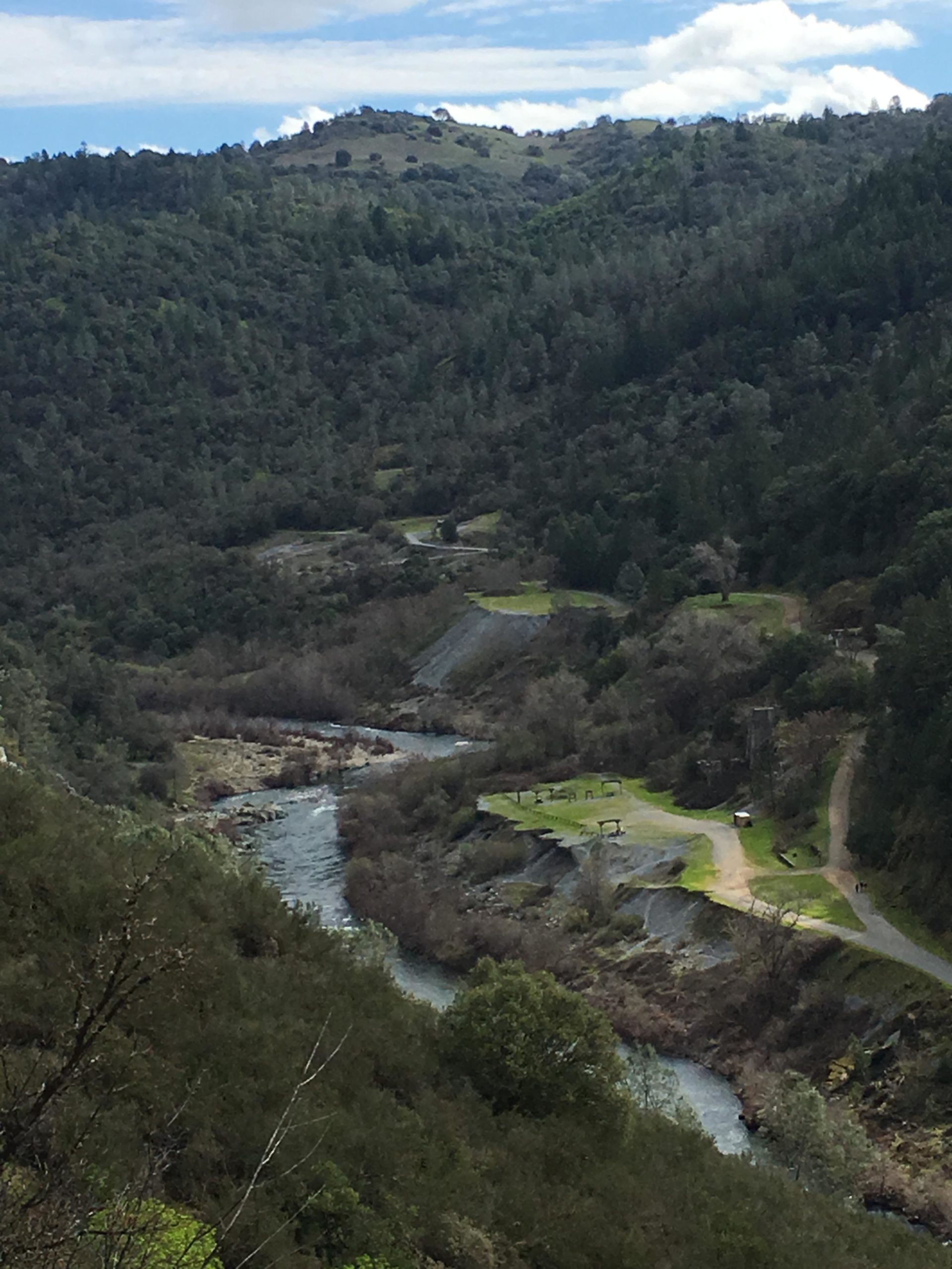A scenic overview of a winding river surrounded by lush green hills and trees, with a walking path and grassy area visible along the riverbank. Clouds are gently scattered in the blue sky above. Foresthill Divide mountain bike trail.