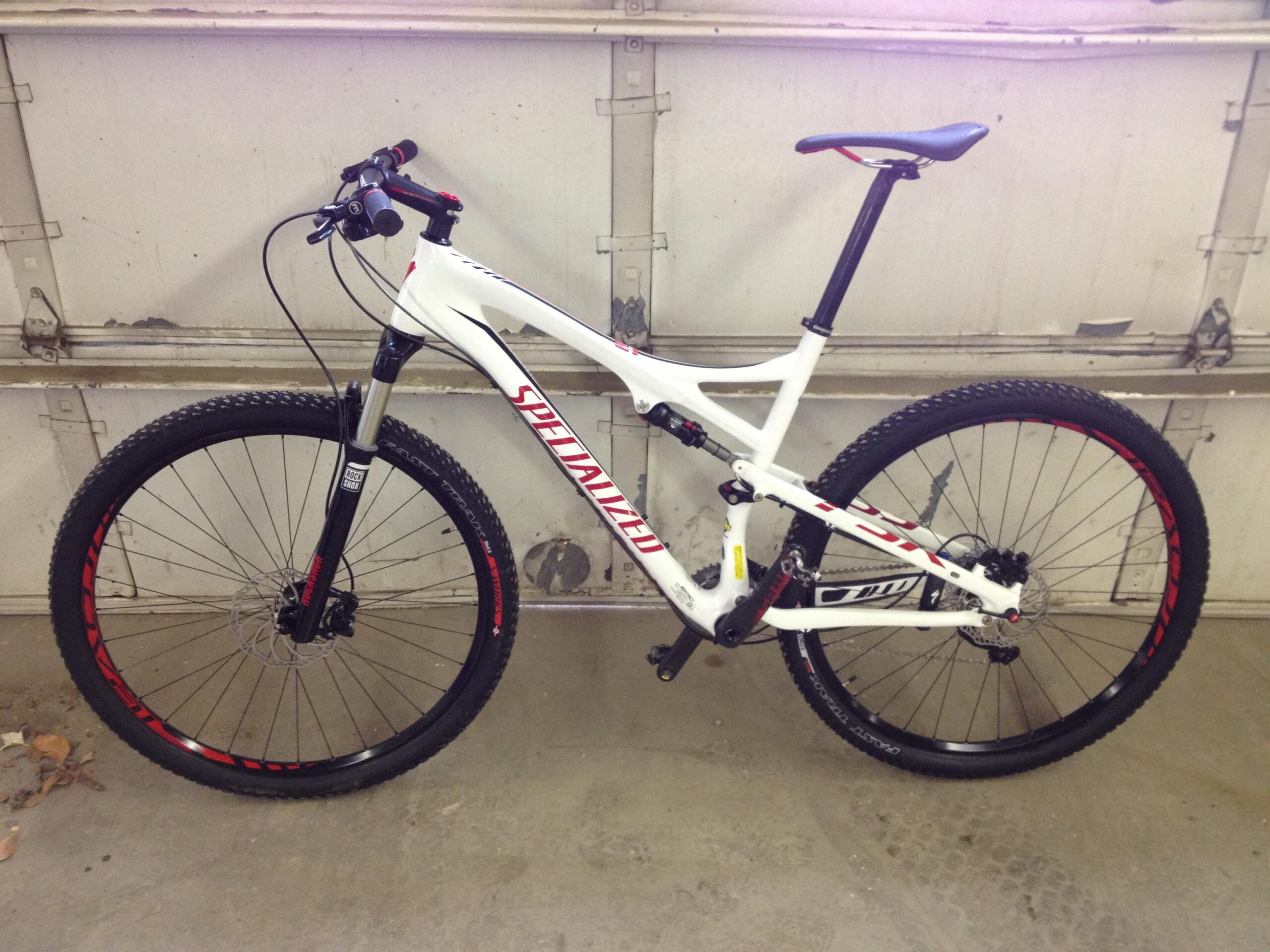 Specialized Epic Expert Carbon 29er: A white mountain bike with red accents leaning against a garage wall. The bike features knobby tires, a suspension fork, and a sleek frame design, making it suitable for off-road riding. The handlebars are equipped with grips and a bell, while the seat is positioned for comfort during rides. The garage floor is concrete, and the background includes a slightly weathered garage door.