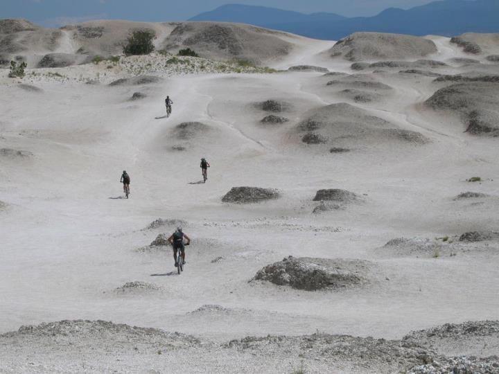 Four mountain bikers riding through a rugged, dusty landscape with white, undulating terrain and small hills under a blue sky. White Ridge Bike Trails mountain bike trail.